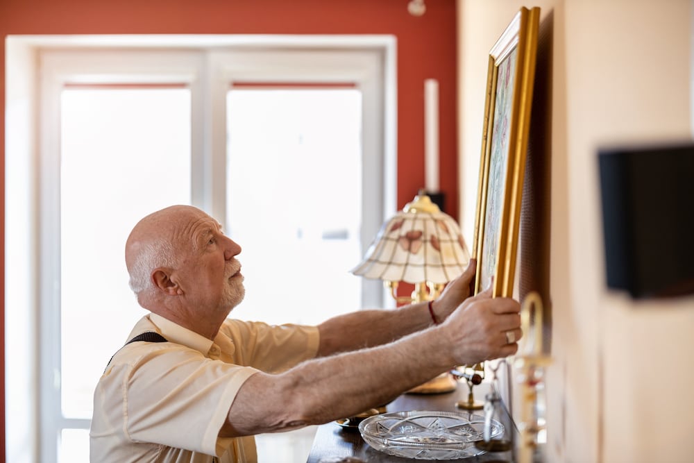 Un anciano colgando un cuadro en su apartamento.